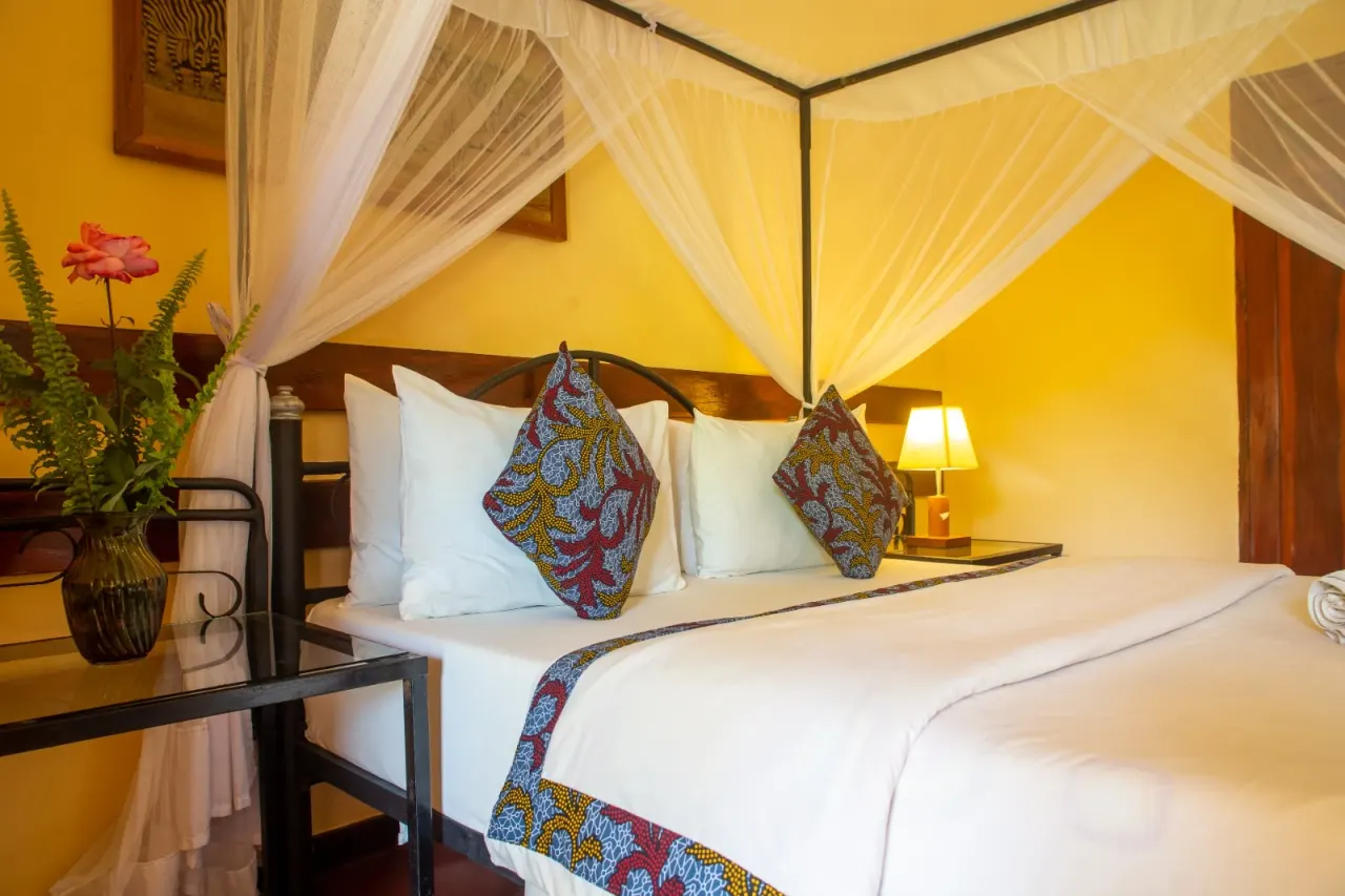 A guest room at Mullers with a four-poster bed under a draped mosquito net, kitenge cushions against a warm yellow wall