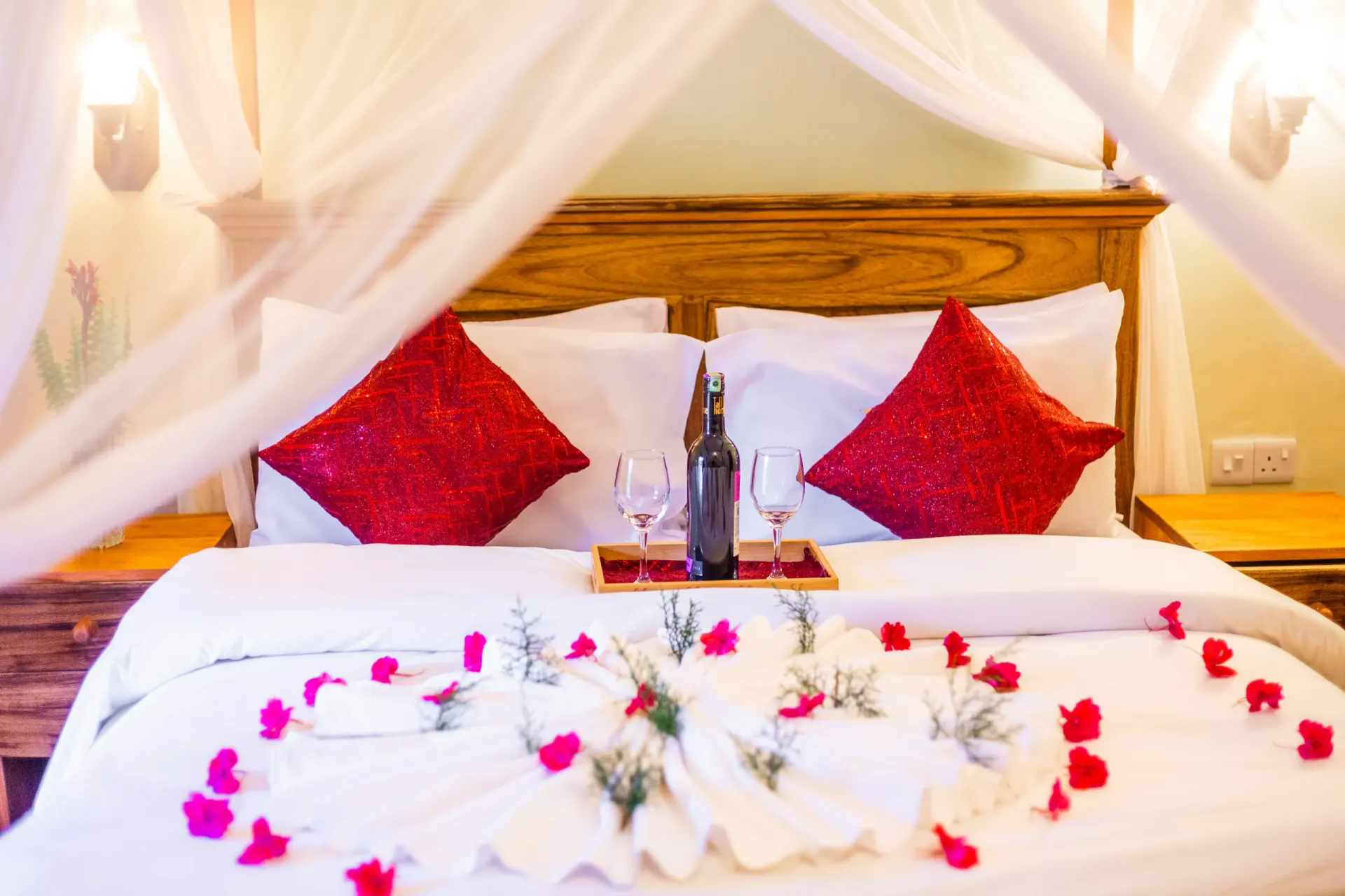 A view room with a wooden four-poster bed under a mosquito net, dressed in white linen and scattered rose petals with wine set for arrival