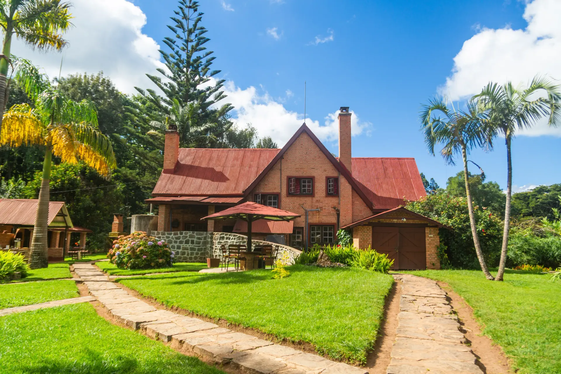 The brick farmhouse with its red roof framed by palms and pines, stone paths crossing the green lawn to a guest cottage